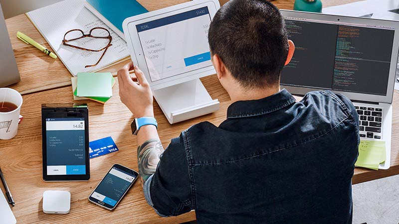 Man with a laptop, tablet, mobile phone, papers and Square payment device on a desk in front of him. Man with a laptop, tablet, mobile phone, papers and Square payment device on a desk in front of him.