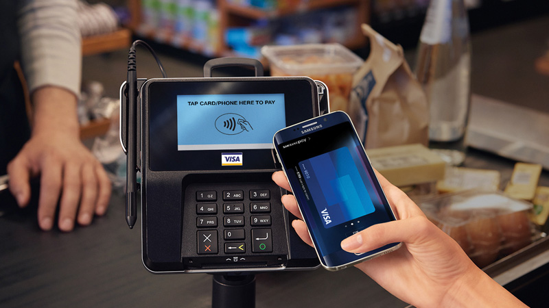 A hand holding a mobile phone showing a Visa card on the screen while in front of a payment terminal. A hand holding a mobile phone showing a Visa card on the screen while in front of a payment terminal.
