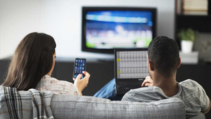 A woman using a smartphone and man using a laptop both sitting on a couch in front of a television. A woman using a smartphone and man using a laptop both sitting on a couch in front of a television.